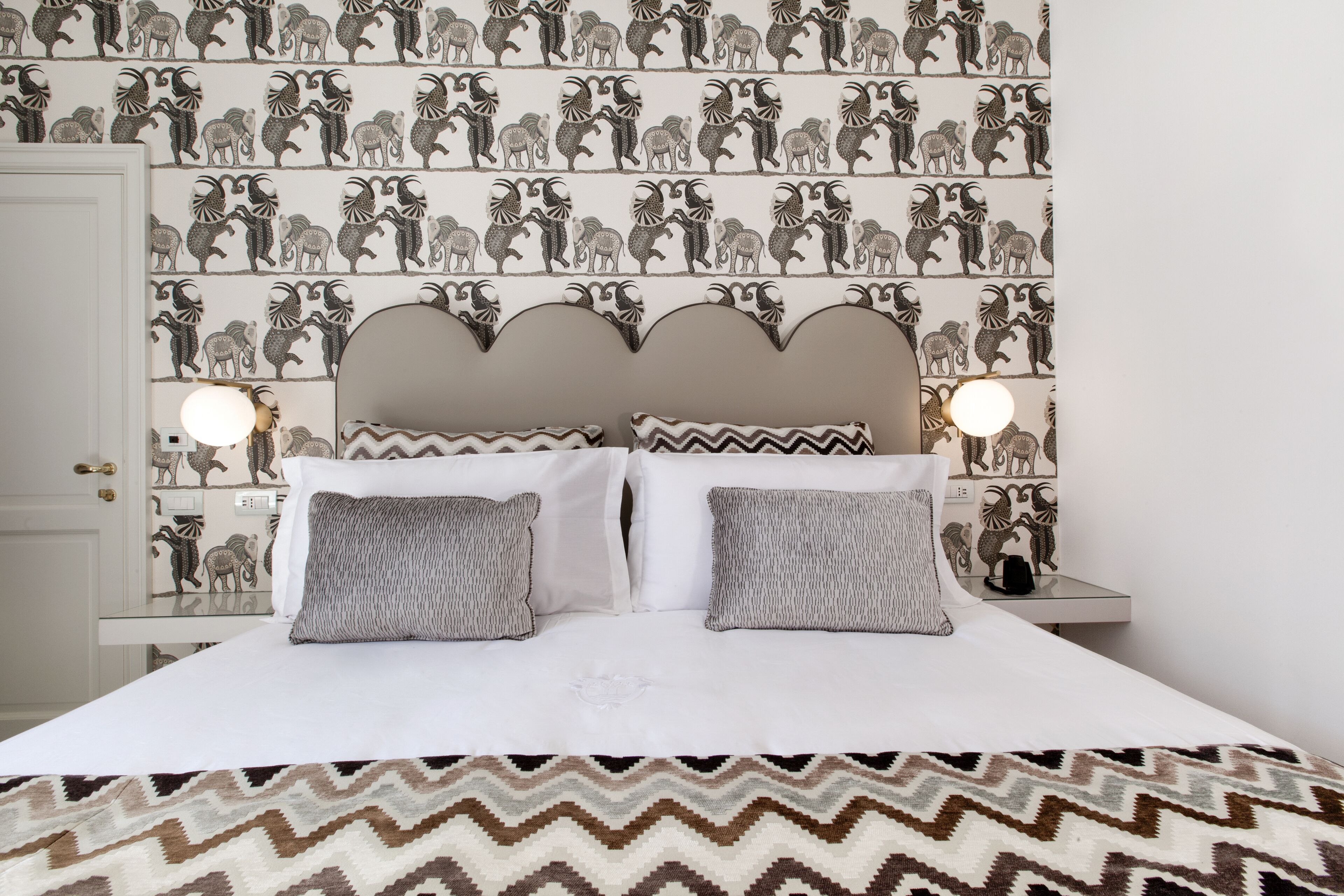 A neatly made bed with patterned pillows and a chevron-patterned bedspread. The room features a wall with a repeating animal print wallpaper and a simple, modern headboard.