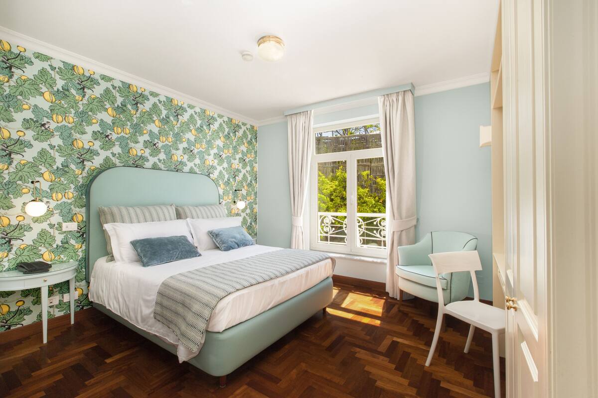 Un dormitorio con cama, mesita de noche, silla y ventana con cortinas.