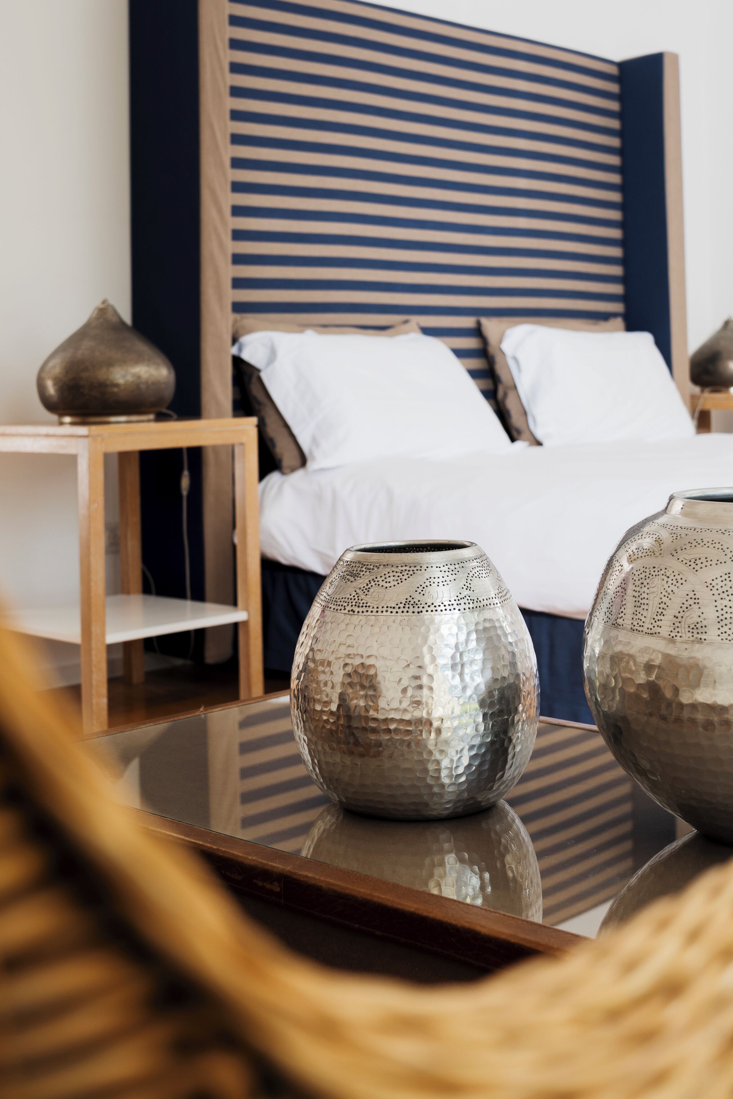A neatly arranged bedroom with a striped headboard, a wooden nightstand, and two metallic vases on a glass table.