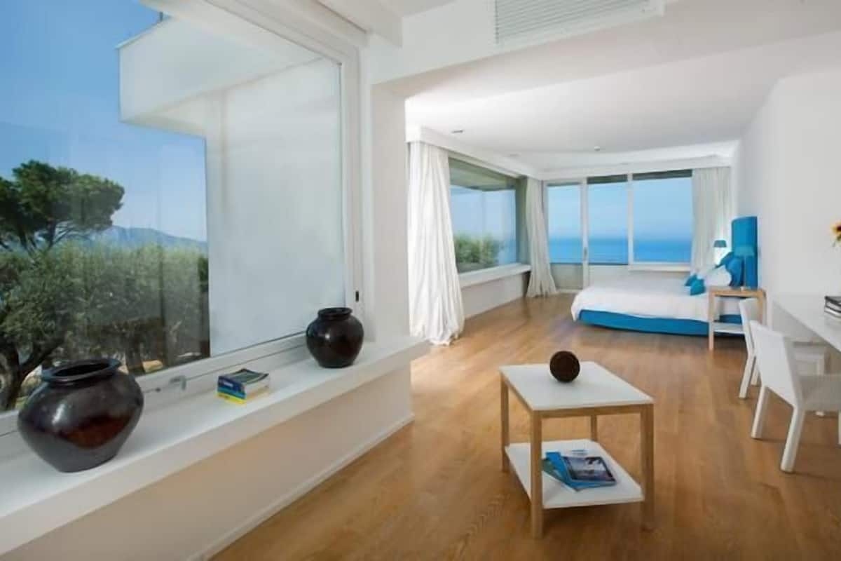 A modern bedroom with a large window, a bed, a desk, and a view of the sea.