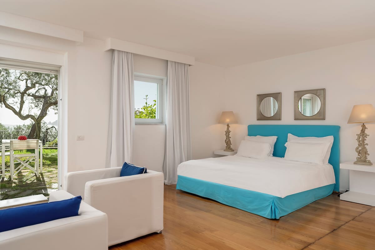 A bedroom with a bed, two bedside tables, a window with curtains, a white sofa, and a view of the outdoors.