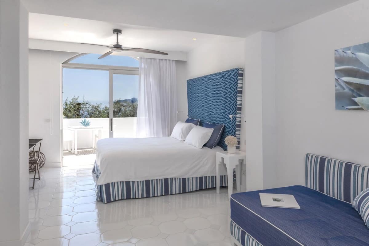 A bedroom with a bed, a bedside table, a chair, a ceiling fan, and a view of the outdoors.