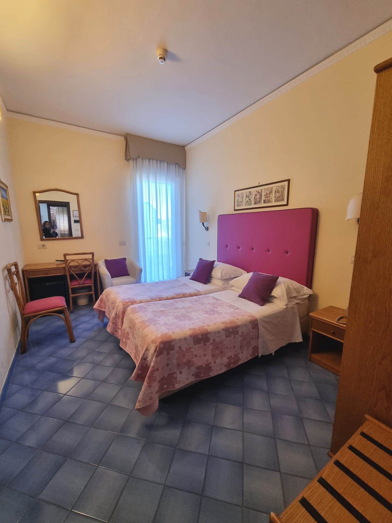 Une chambre avec deux lits, un bureau, une chaise, un miroir et une fenêtre avec des rideaux.