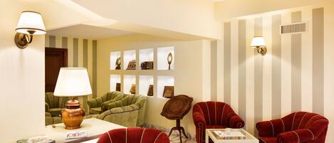 Habitación de hotel con paredes a rayas, un sofá verde, sillones rojos, una mesita y lámparas fijadas a la pared.