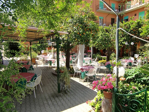 Un patio al aire libre con mesas y sillas, rodeado de follaje y plantas en macetas.
