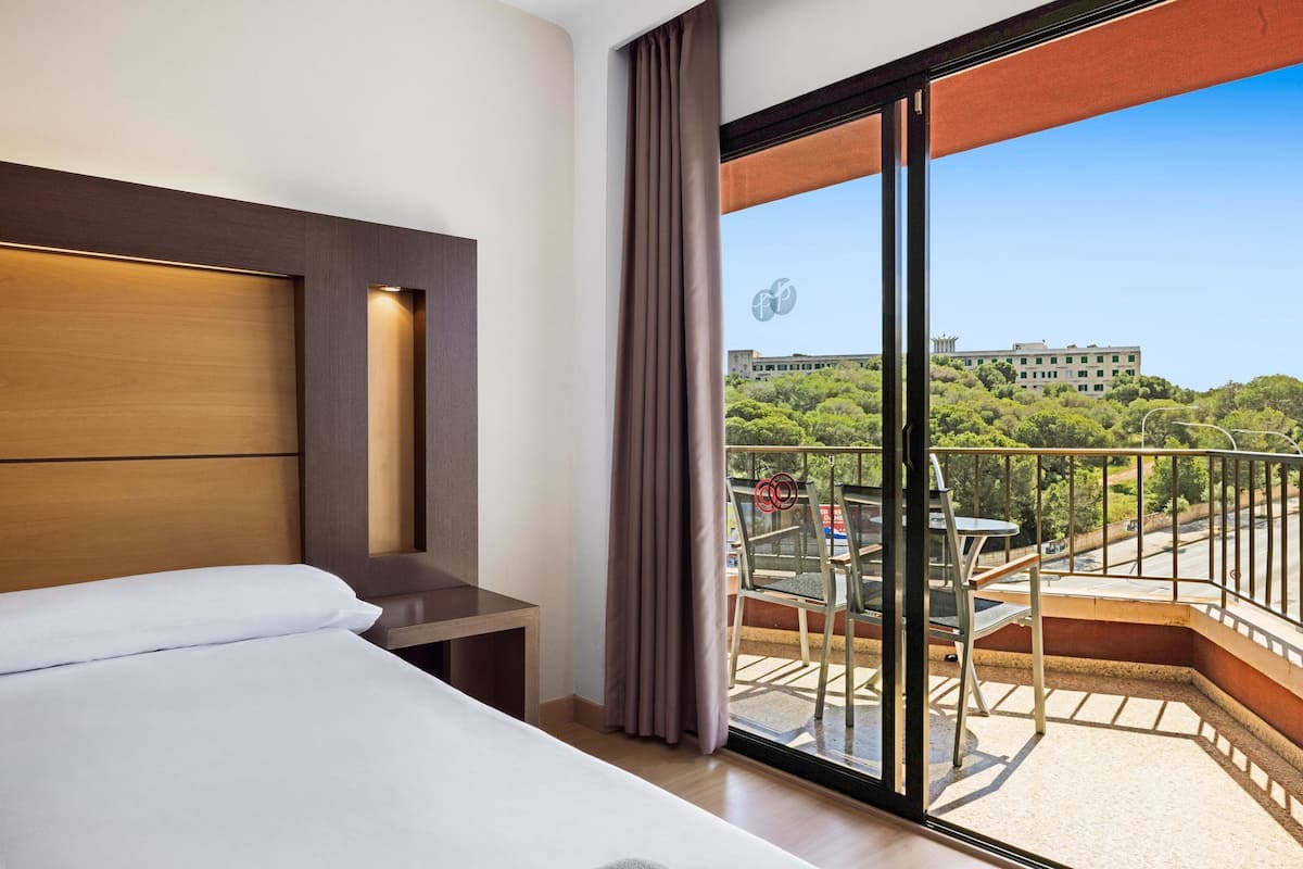 Habitación de hotel con cama, escritorio, balcón con mesa y sillas, y vista a un edificio y árboles.