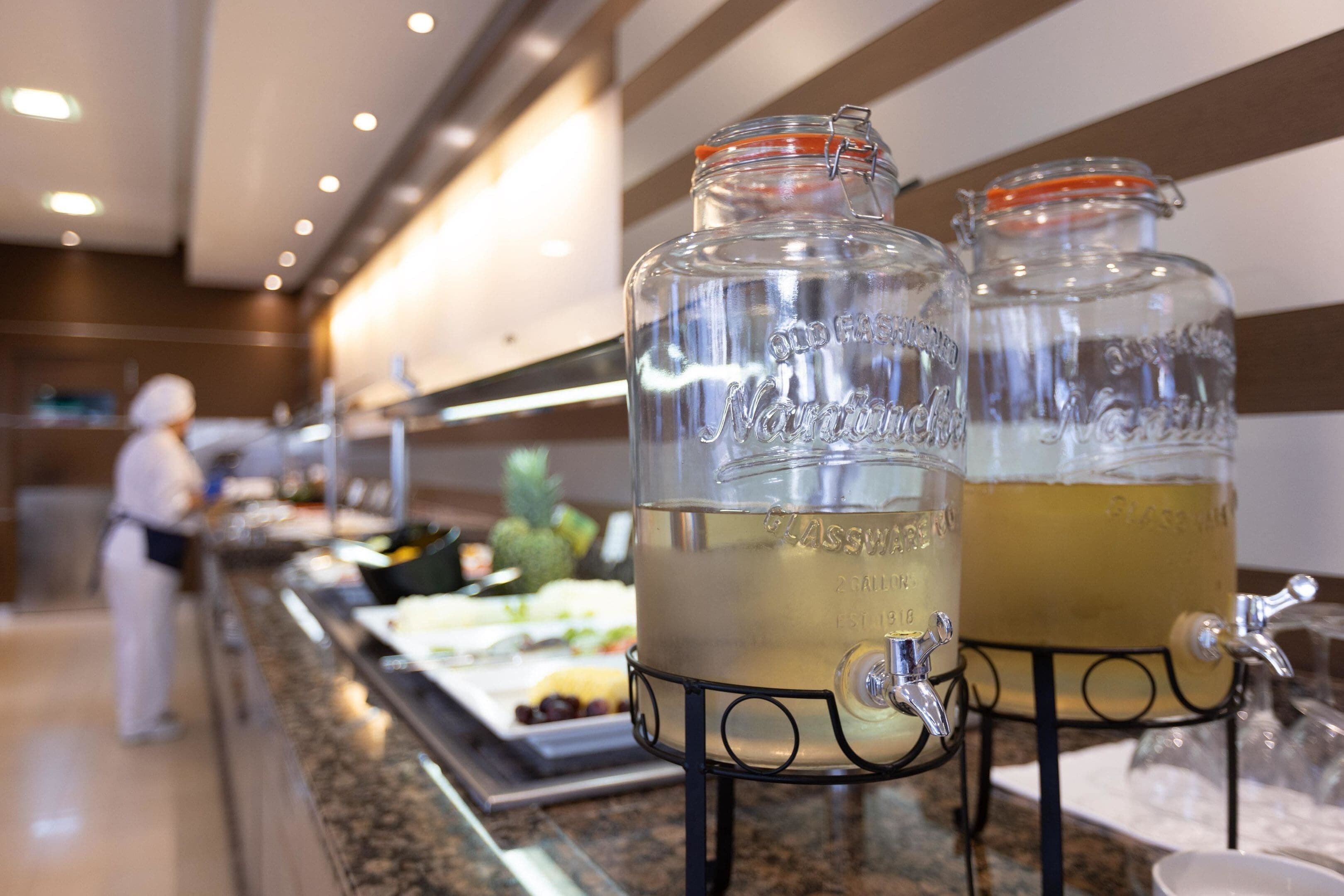 Two glass dispensers with metal taps on a countertop, a buffet line with food and drinks, and a person in the background.