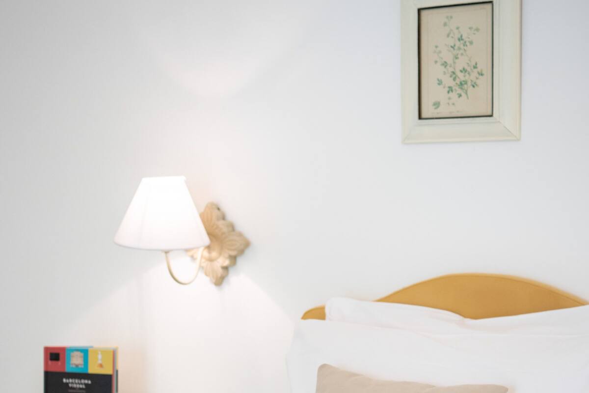 A neatly arranged hotel room with a bed, a bedside table with a lamp, a framed picture on the wall, and a small box of items on the table.