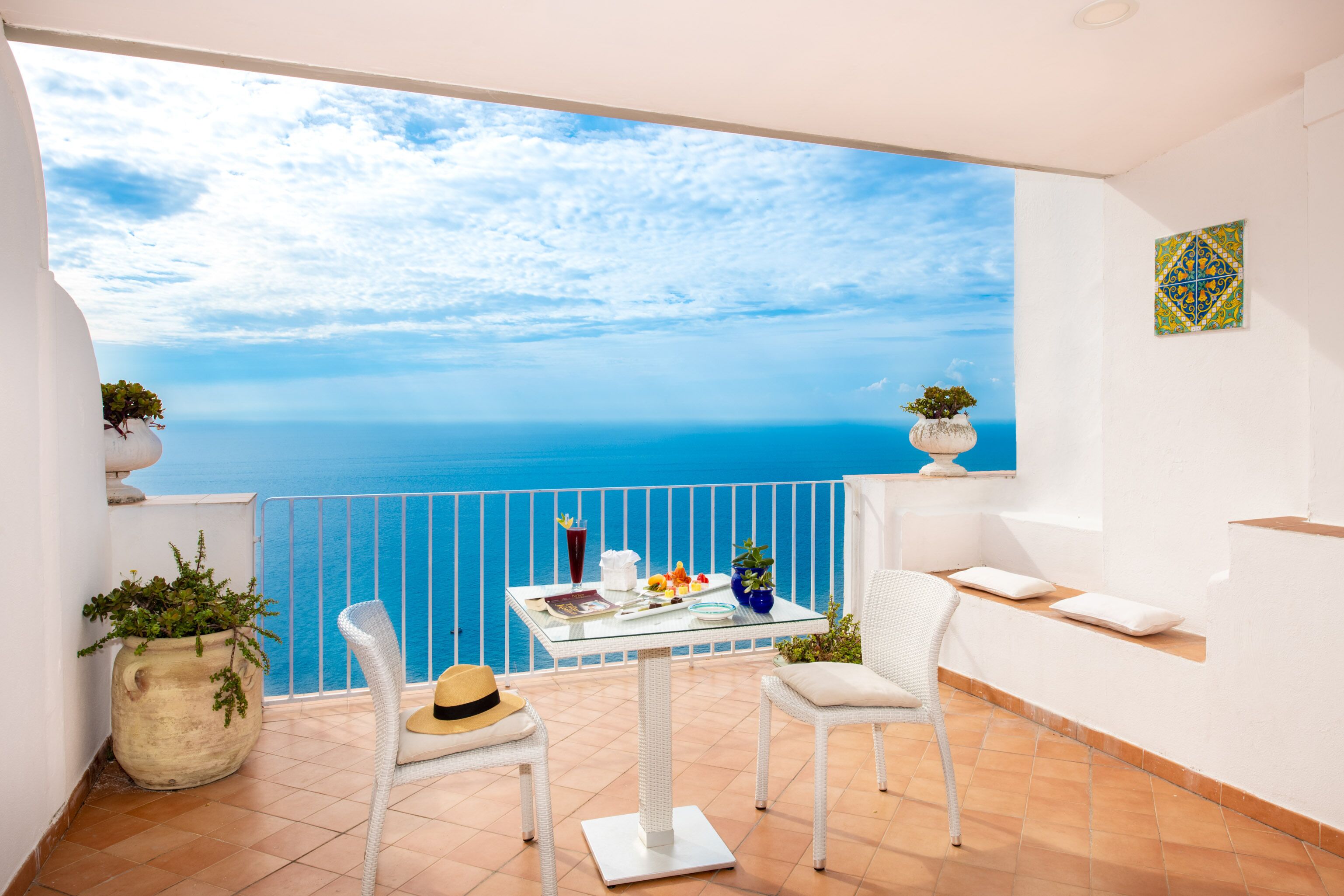 A balcony with a glass table, two white wicker chairs, a potted plant, and a view of the sea.