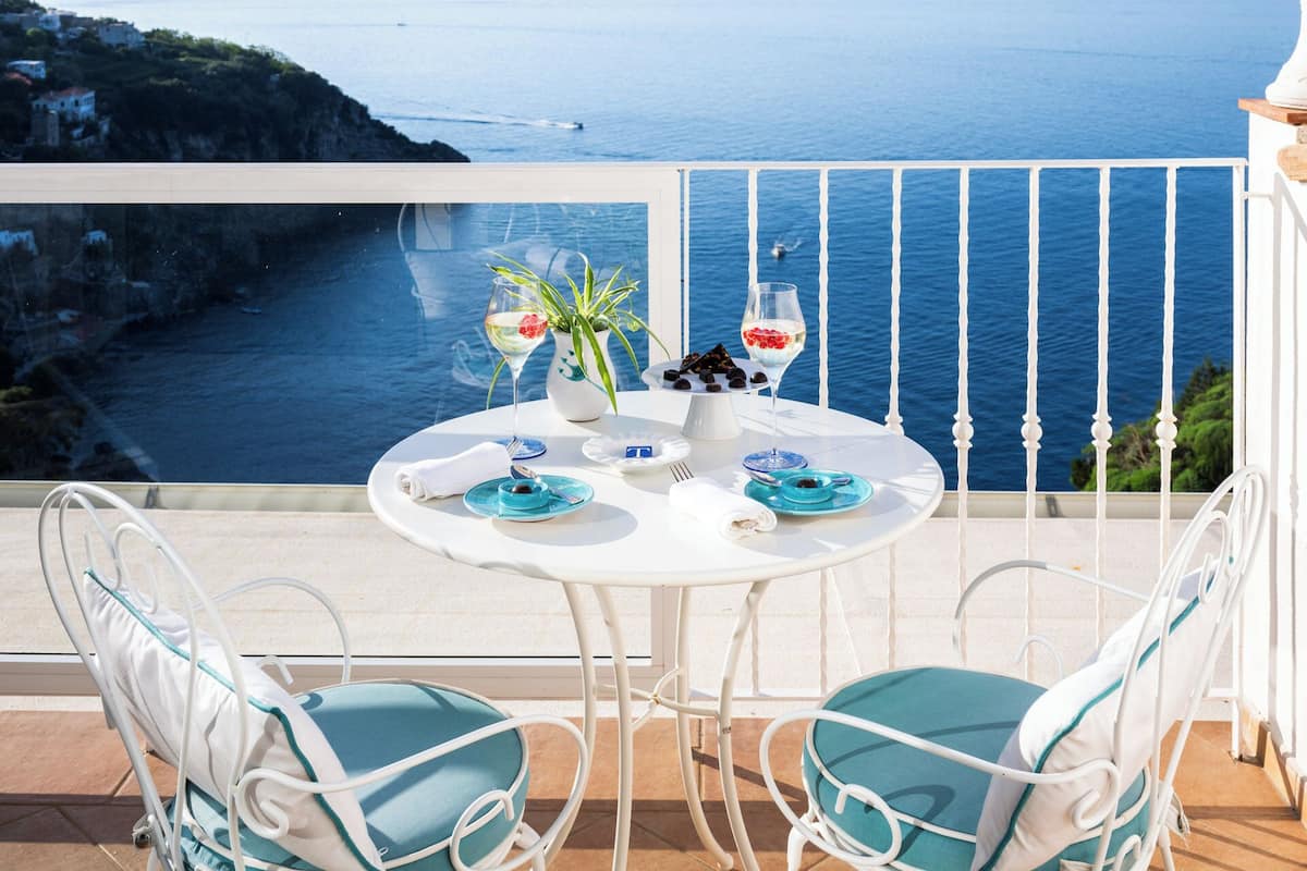 A balcony with a white table set for two, overlooking a coastal view.