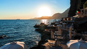 Un resort costero con aguas iluminadas por el sol, un acantilado rocoso y mesas al aire libre.