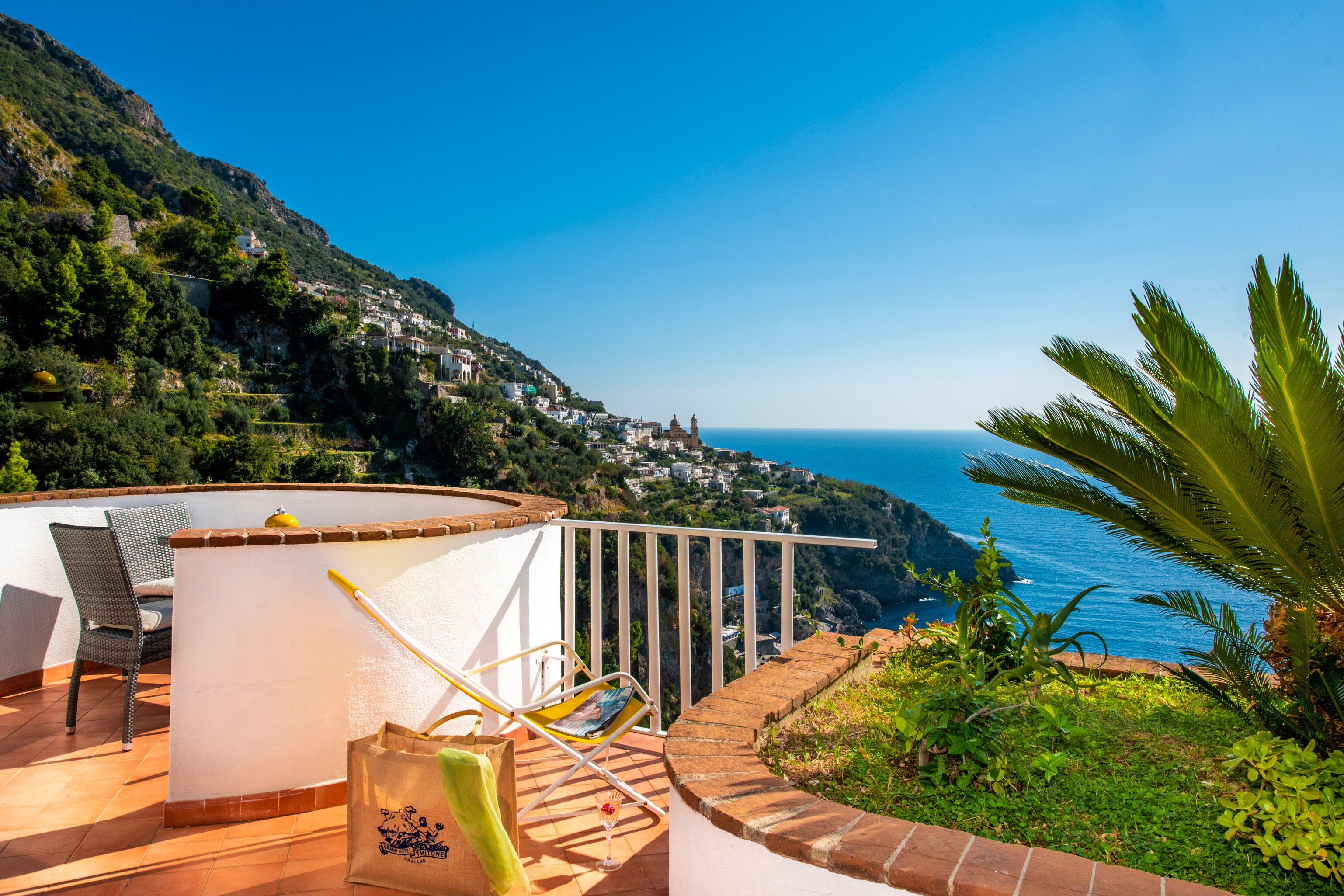 A terrace with a white railing, a chair, a table, and a view of a coastal town and ocean.