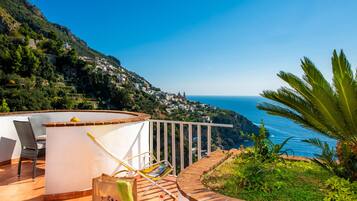 A terrace with a white railing, a chair, a table, and a view of a coastal town and ocean.