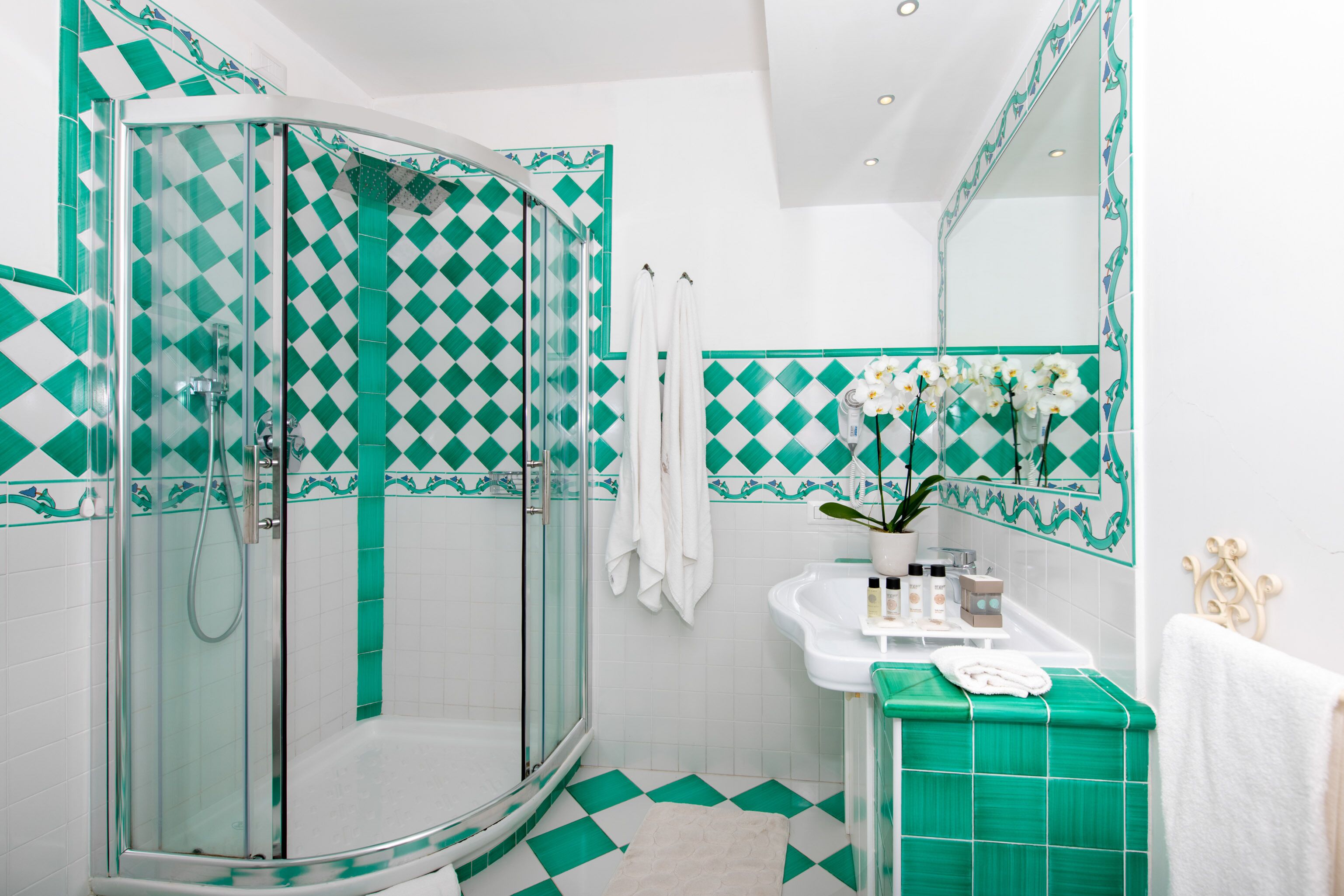 A bathroom with a glass-enclosed shower, a sink with a mirror, and a tiled floor with a green and white pattern.