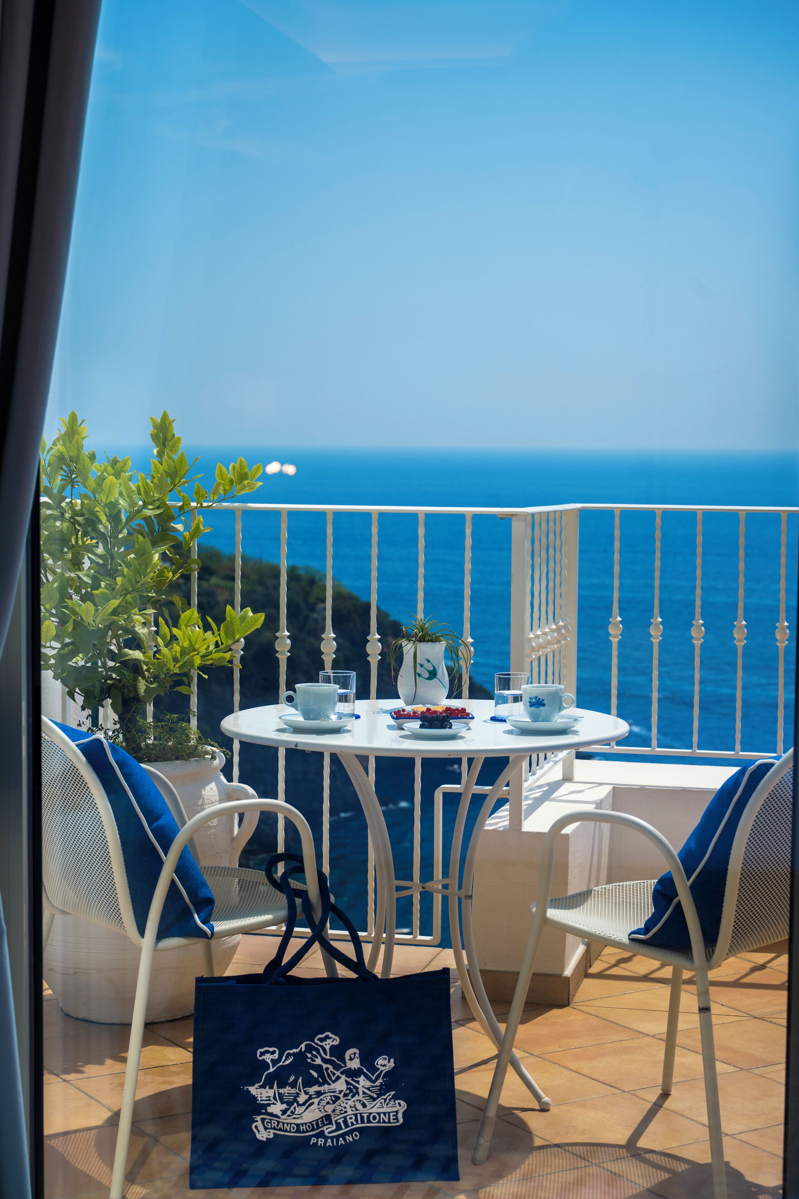 A balcony with a white table and chairs, a potted plant, and a view of the sea.