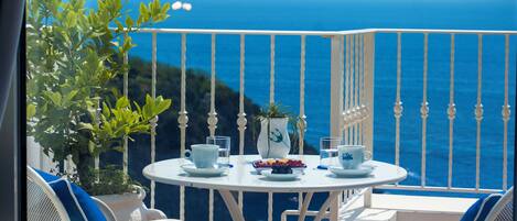A balcony with a white table and chairs, a potted plant, and a view of the sea.