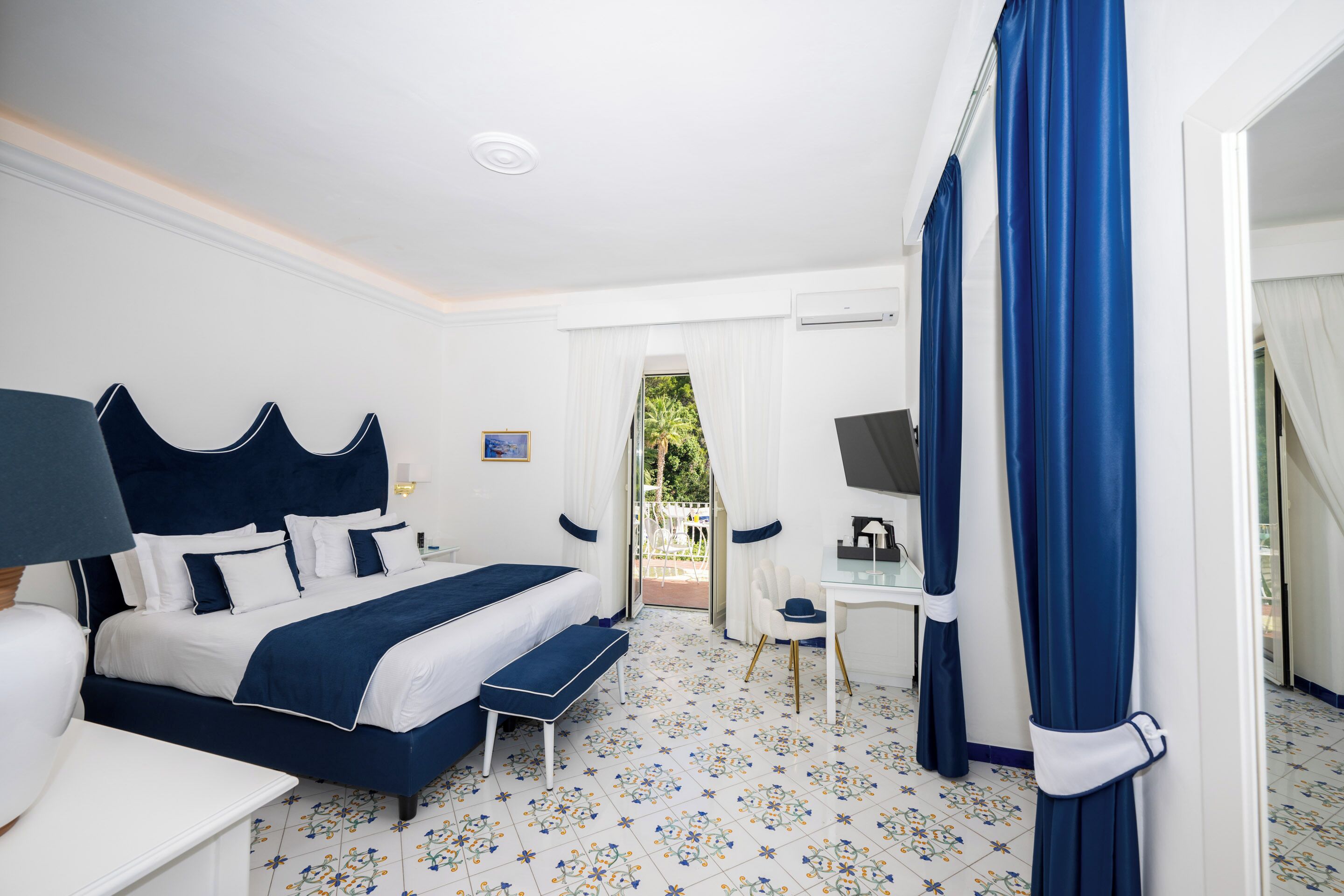 A hotel room with a large bed, a desk, and a chair. There is a window with curtains and a view of the outdoors.