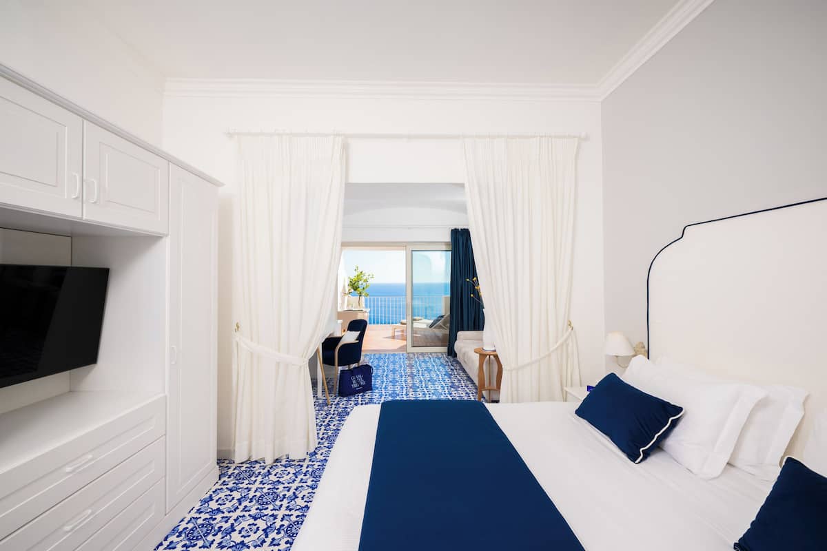 A hotel room with a bed, a TV, a desk, and a balcony with a view of the sea.