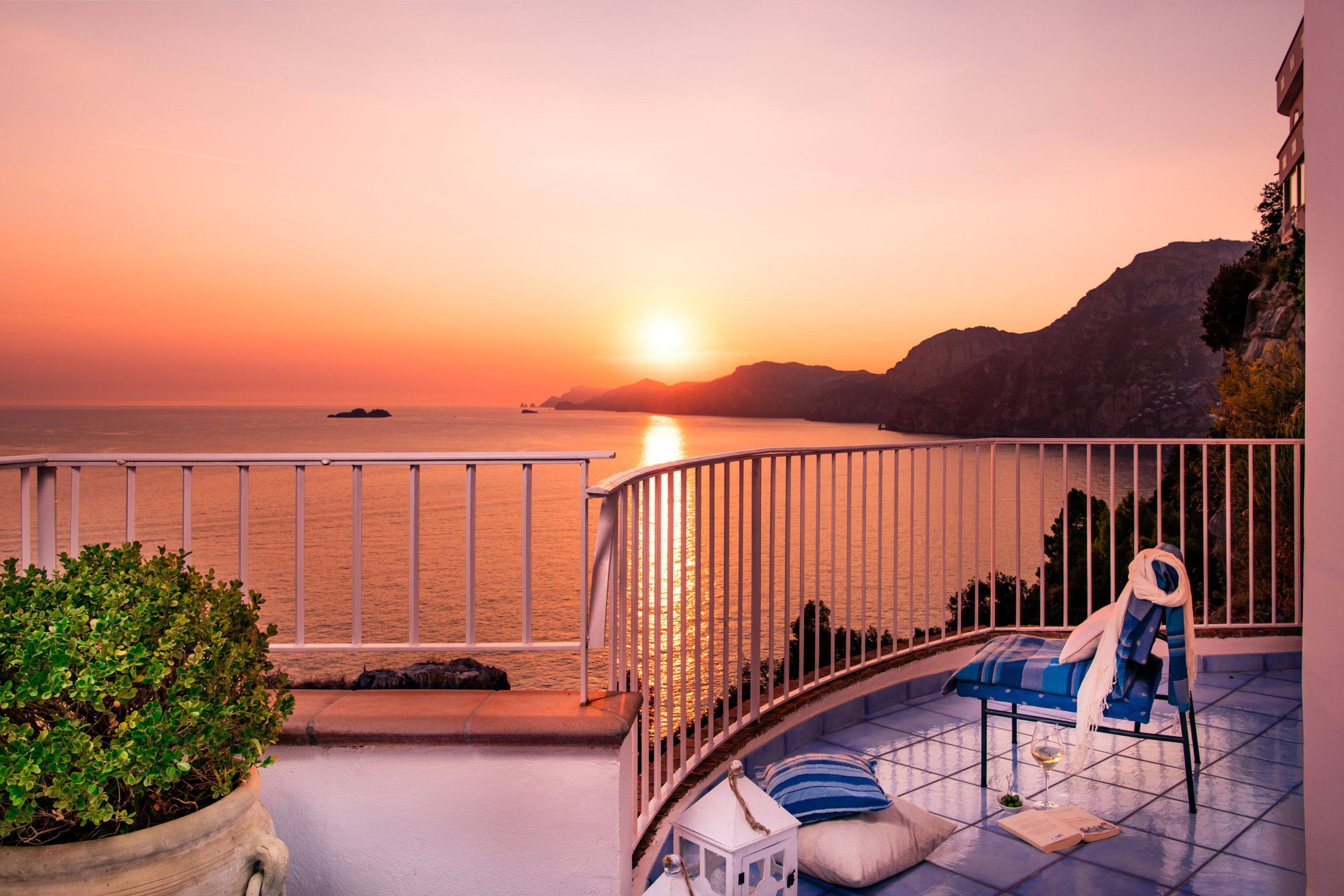 A balcony with a view of the sunset over the sea, a potted plant, and a lounge chair with a towel.
