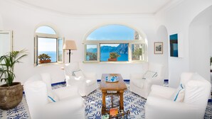 A spacious room with white furniture, a wooden coffee table, and large windows offering a view of the sea and mountains. - Grand Hotel Tritone (Praiano)