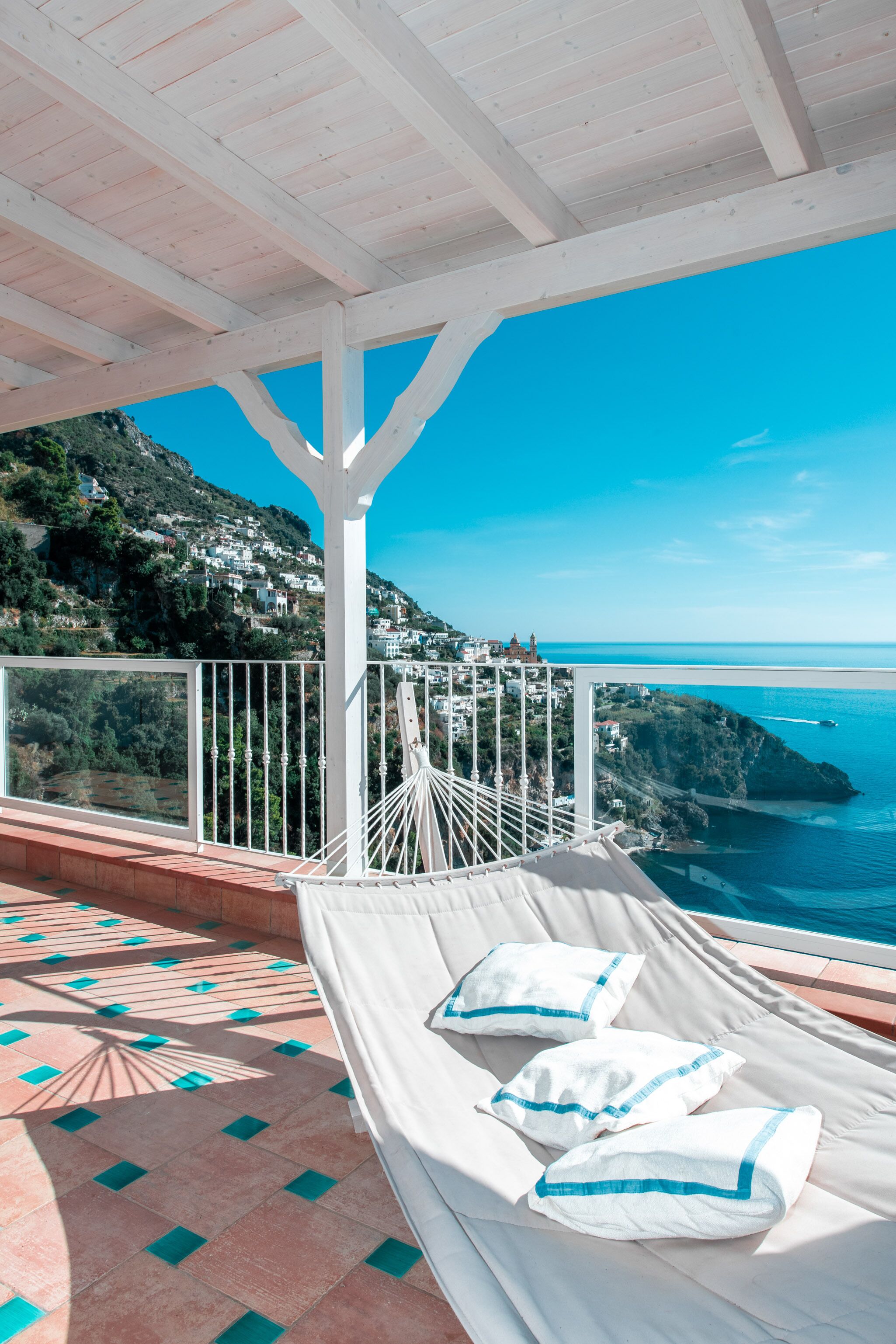 A balcony with a white cushioned bench, white railings, and a view of the sea and hills.