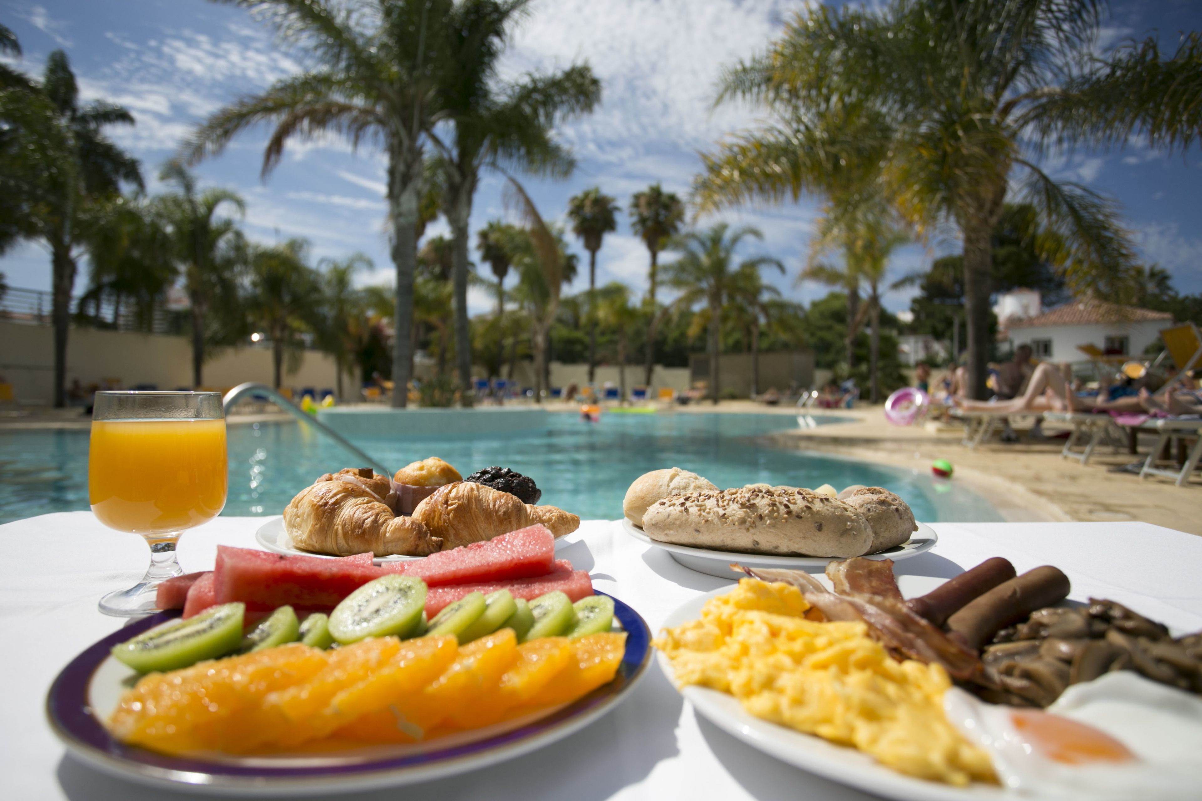 A breakfast table with croissants, fruit, and eggs by a poolside with palm trees.