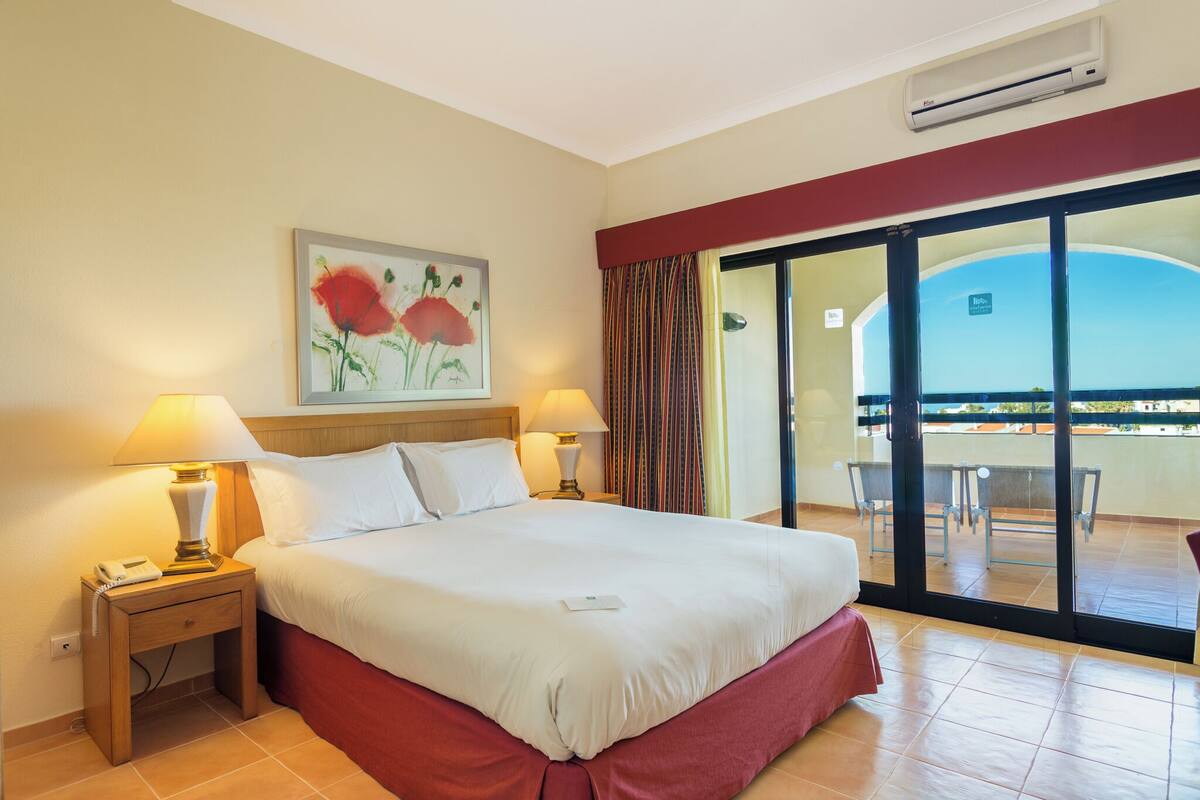 Una habitación de hotel con cama, dos lámparas de noche, un cuadro, aire acondicionado y un balcón con vistas.