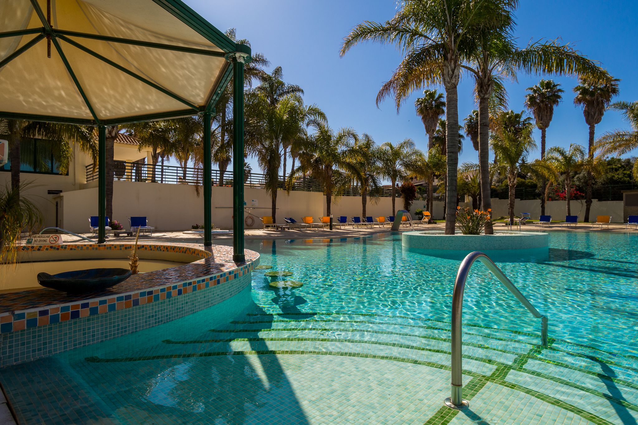 A swimming pool with tiled edges, lounge chairs, palm trees, and a covered area.