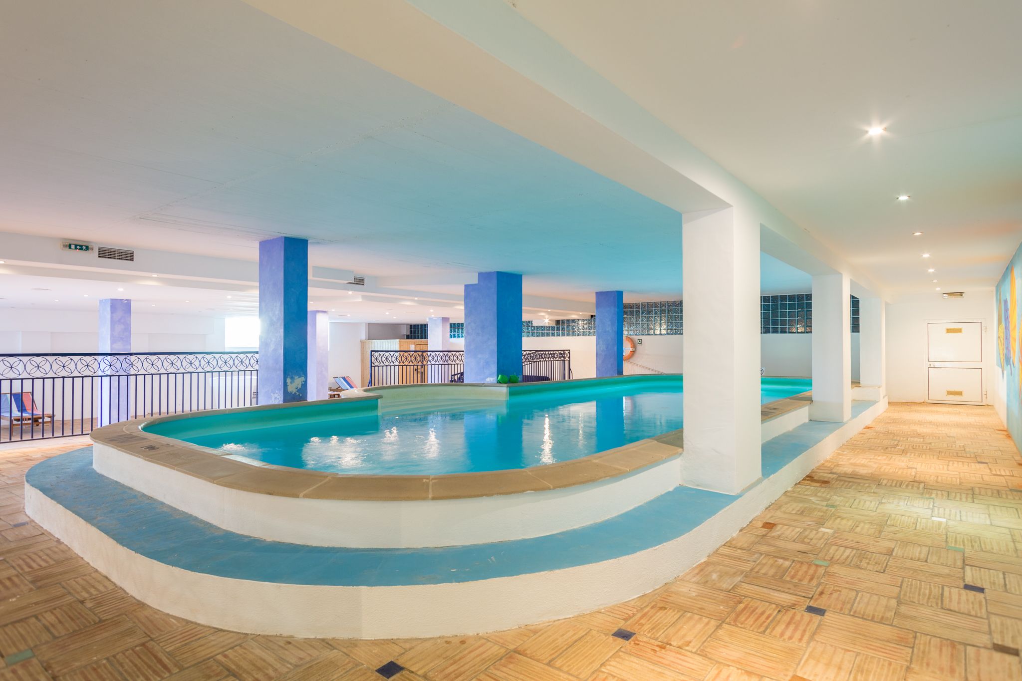 An indoor swimming pool with a tiled floor, surrounded by white pillars and a railing.