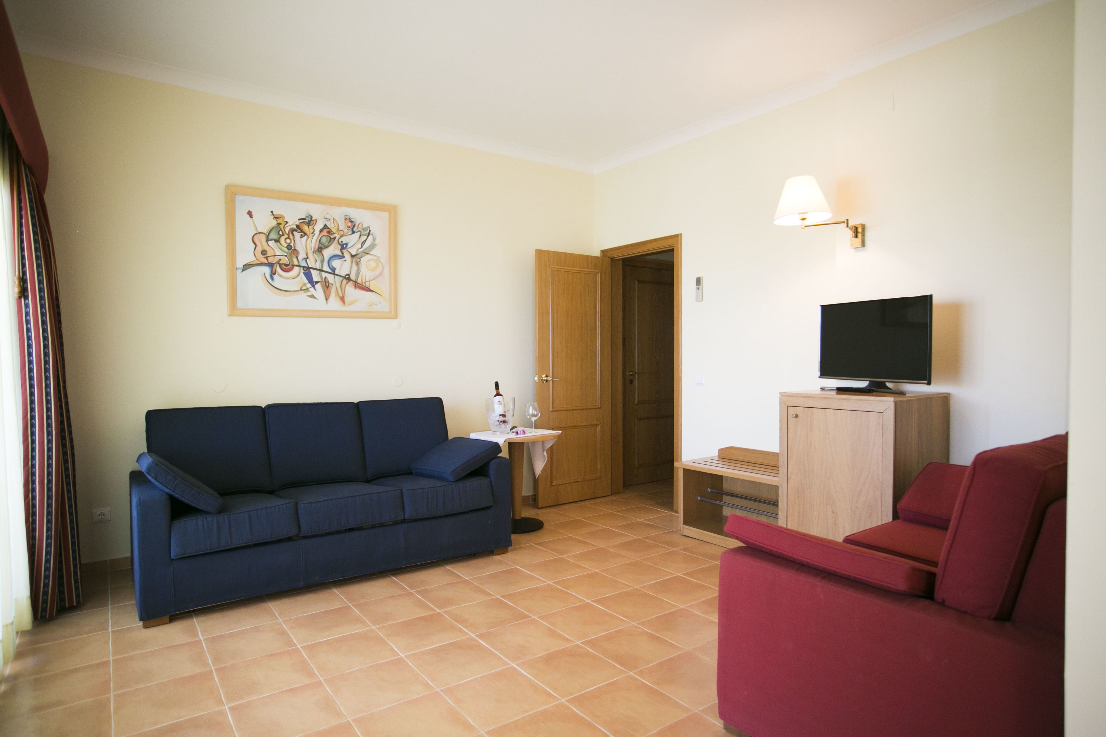 A living room with a blue sofa, a red armchair, a television on a wooden cabinet, a small table with a wine bottle, and a door leading to another room.