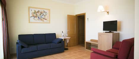 A living room with a blue sofa, a red armchair, a television on a wooden cabinet, a small table with a wine bottle, and a door leading to another room.