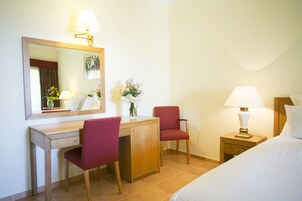 Habitación de hotel con cama, un escritorio con silla, una mesita de noche con lámpara, un espejo y un jarrón con flores.