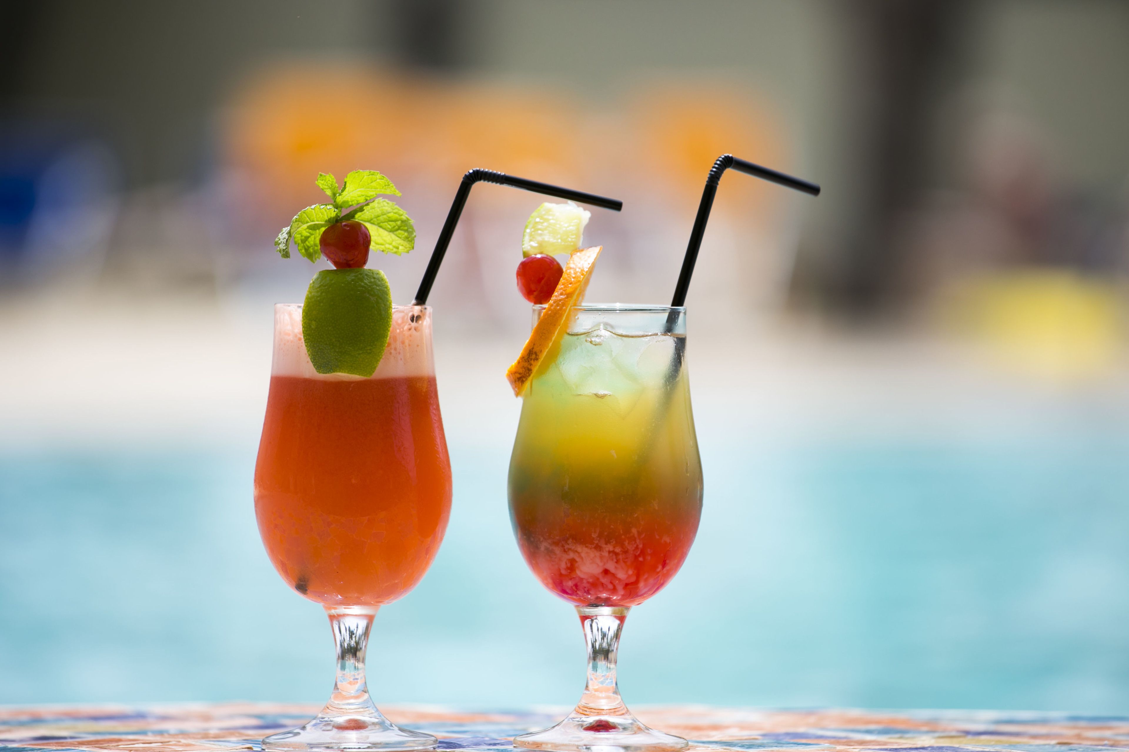 Two cocktails with straws and garnishes, served in stemmed glasses, on a table by a pool.