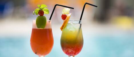 Two cocktails with straws and garnishes, served in stemmed glasses, on a table by a pool.