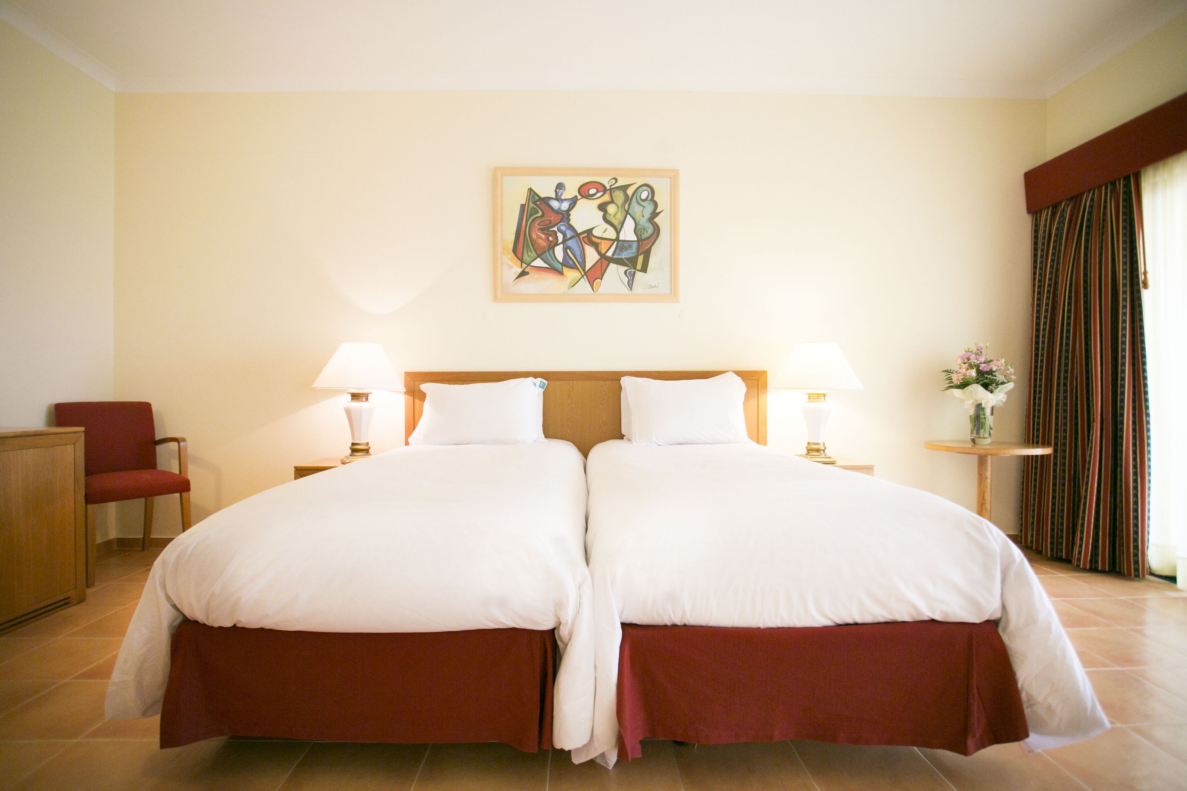 Une chambre d’hôtel avec deux lits, une chaise, une lampe, un tableau au mur et une petite table sur laquelle se trouve un vase.