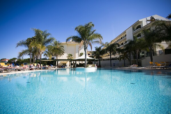A swimming pool with lounge chairs and palm trees, surrounded by a building with multiple balconies. - Mirachoro Carvoeiro (Lagoa)
