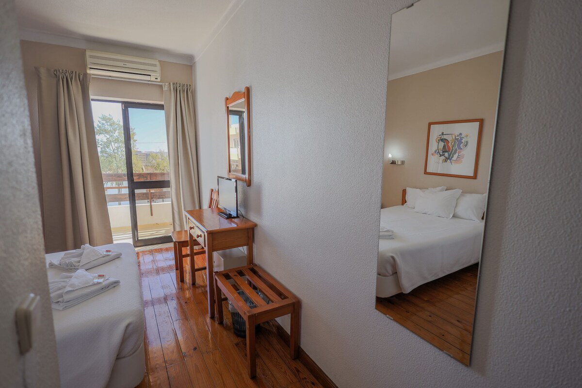 A hotel room with a bed, a desk, a chair, a small table, a mirror, and a window with curtains.