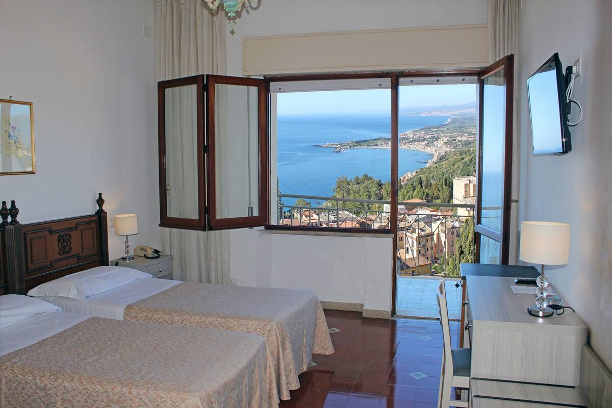 A hotel room with two beds, a desk, a chair, a window with a view of the sea and a town, and a lamp on the desk.
