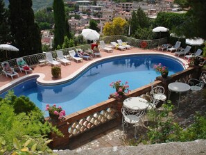 A swimming pool with lounge chairs and umbrellas, surrounded by greenery and a view of a town. - Hotel Mediterranee (Taormina)