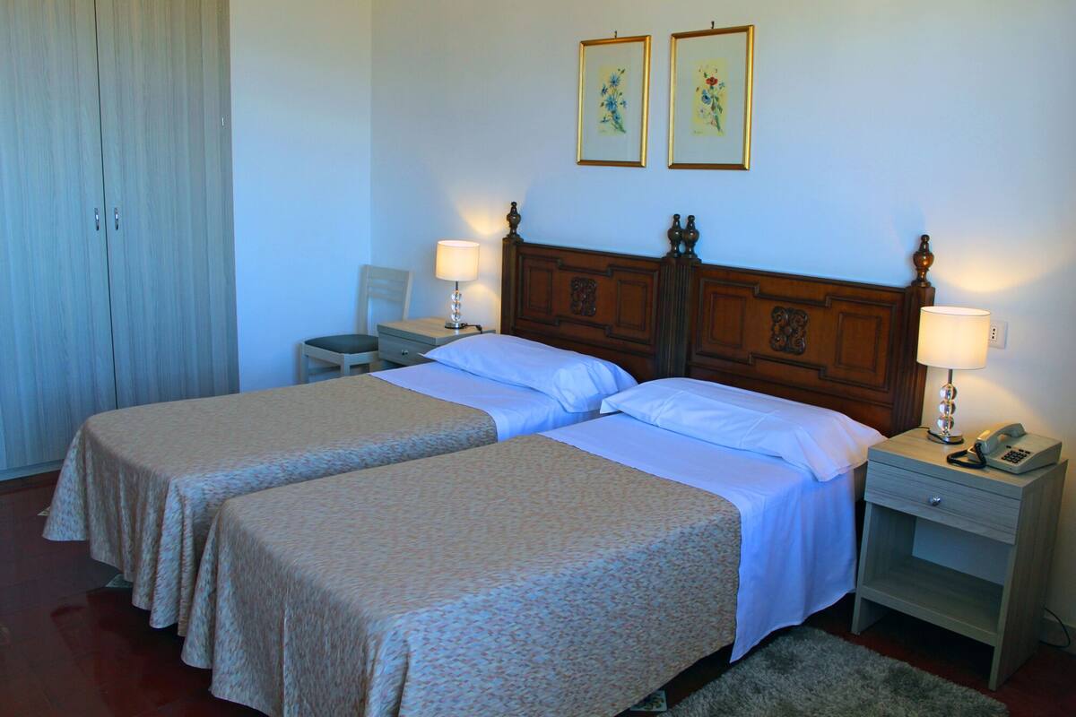 A double room with two beds, a wooden headboard, bedside tables with lamps, a chair, and a wardrobe.