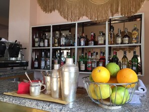 A kitchen counter with a fruit bowl containing oranges and apples, a coffee maker, and a shelf with various bottles of alcohol. - Hotel Mediterranee (Taormina)
