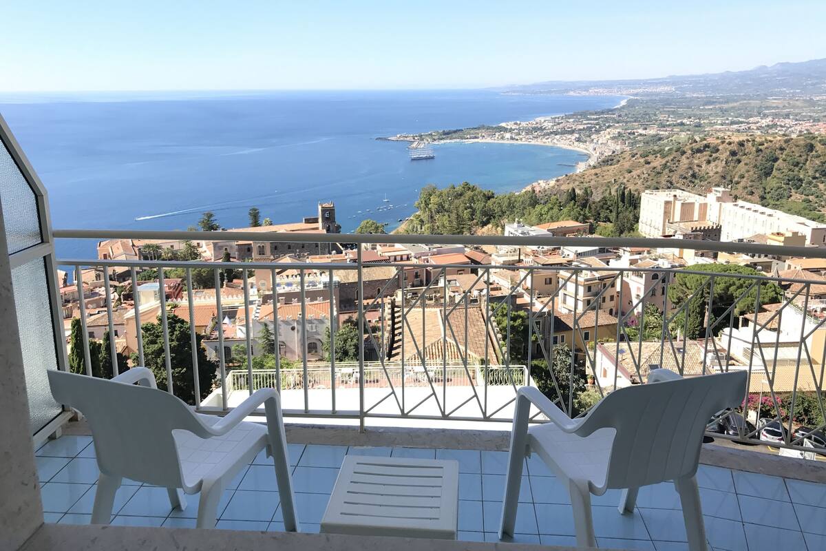 A balcony with two white chairs, a tiled floor, and a view of the sea and buildings.