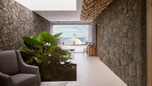 Un intérieur moderne avec un mur en pierre, une grande plante, un canapé et une vue sur l’océan par de grandes fenêtres.