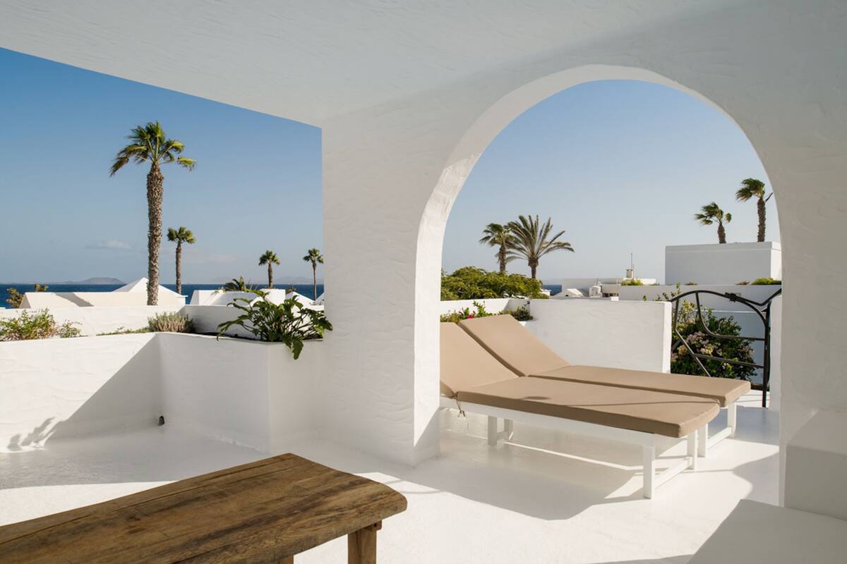 Ein moderner Außenbereich zum Entspannen mit einem Holztisch, weißen Wänden und Blick auf das Meer und Palmen.