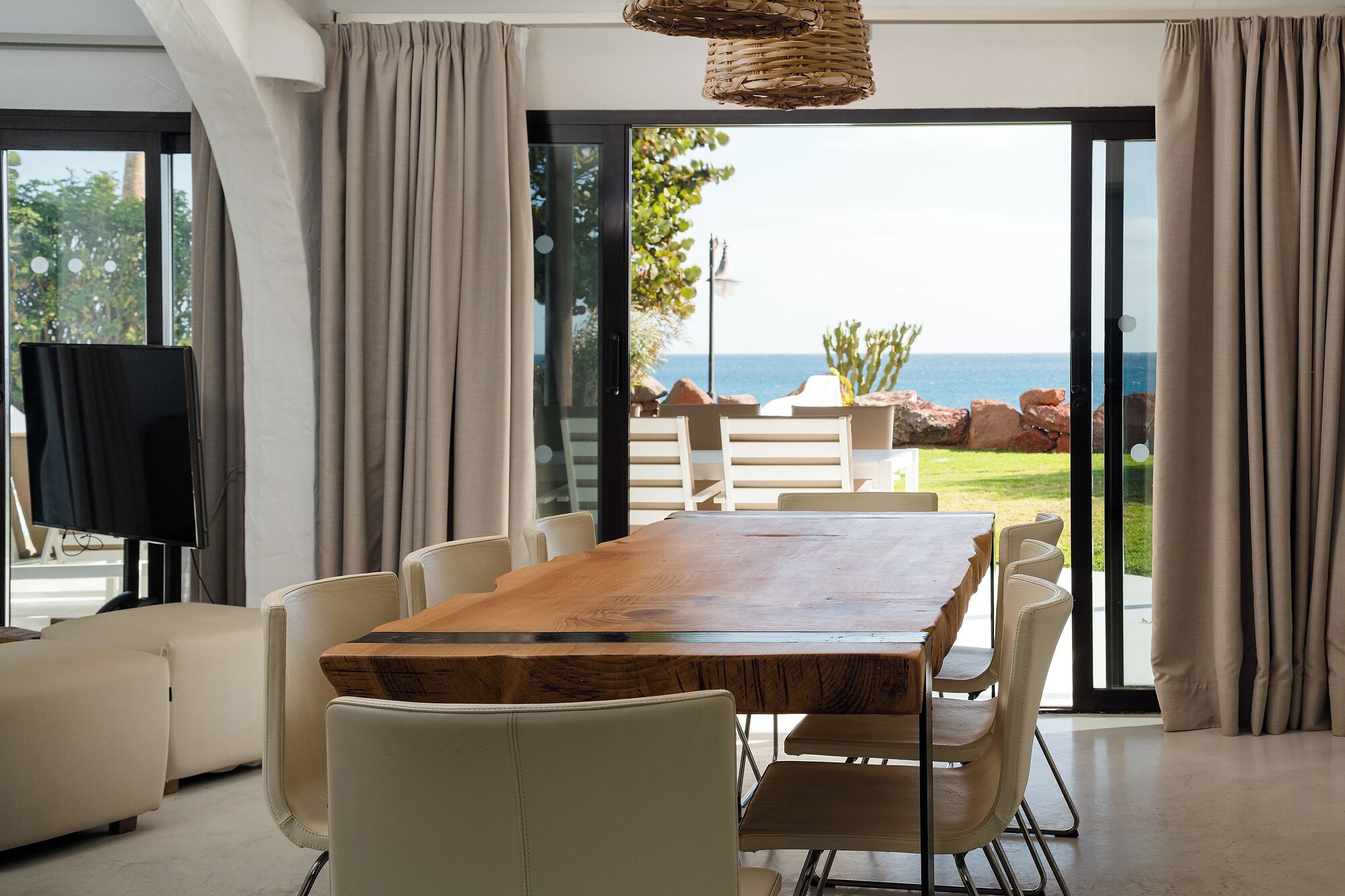 A modern dining area with a wooden table and white chairs, large windows with curtains, and a view of the sea.