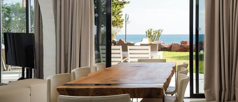 A modern dining area with a wooden table and white chairs, large windows with curtains, and a view of the sea.