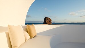 Une chambre donnant sur la plage, avec un canapé blanc et des coussins, un grand rocher en arrière-plan et une vue sur la mer et les montagnes lointaines.