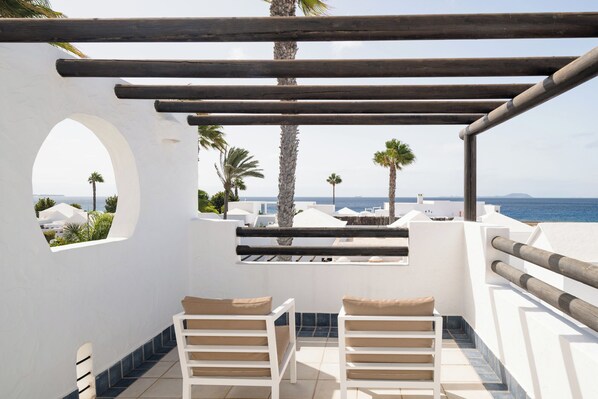Una terraza con paredes blancas, una pérgola de madera, dos sillones blancos con cojines beige, y vistas al mar y a las palmeras.