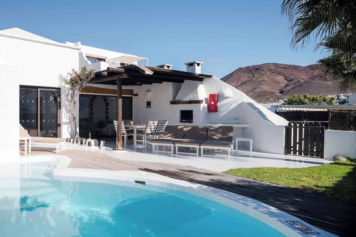 Ein moderner Poolbereich im Freien mit Liegestühlen und einer Pergola, umgeben von einem weißen Gebäude mit Blick auf die Berge.