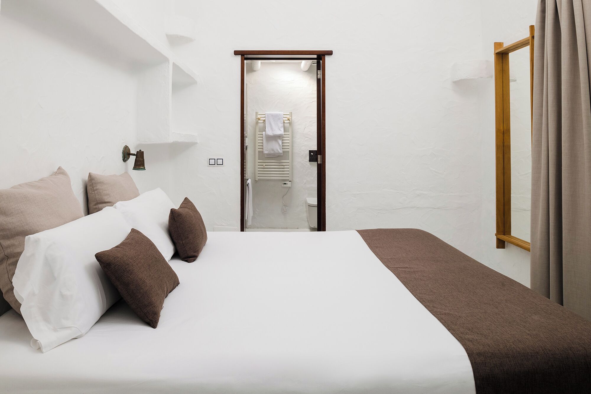 Un dormitori amb un llit, almoines, una tauleta de nit, una porta de fusta i una finestra amb cortines.